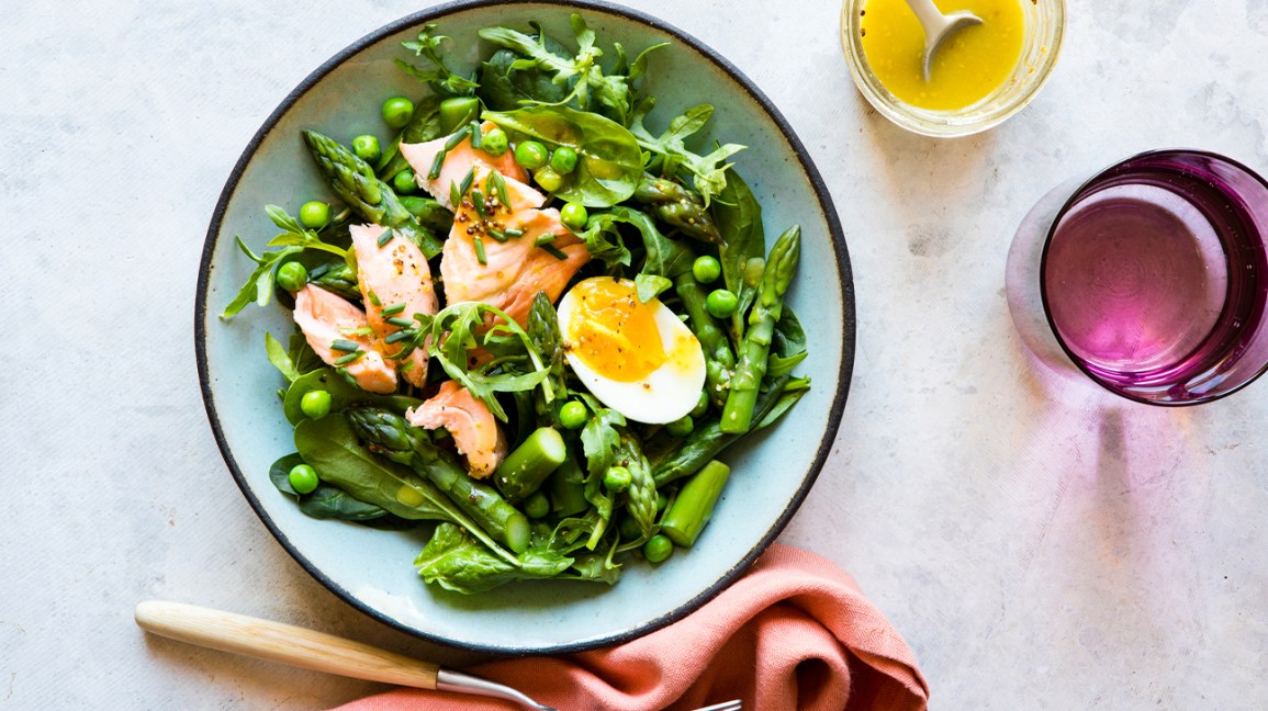 salada feita de espinafre, salmão, ovo, ervilhas e aspargos ao lado de um garfo, guardanapo de pano e copos de molho para salada e água